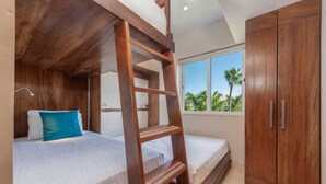 Ein Zimmer mit Etagenbetten, einer Holzleiter, einem Kleiderschrank und einem Fenster mit Blick auf Palmen.