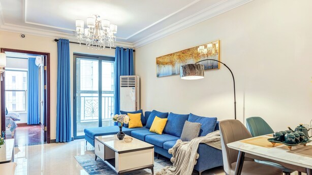 Een moderne woonkamer met een blauwe bank, een salontafel, een eettafel met stoelen, een kroonluchter en een schilderij aan de muur.