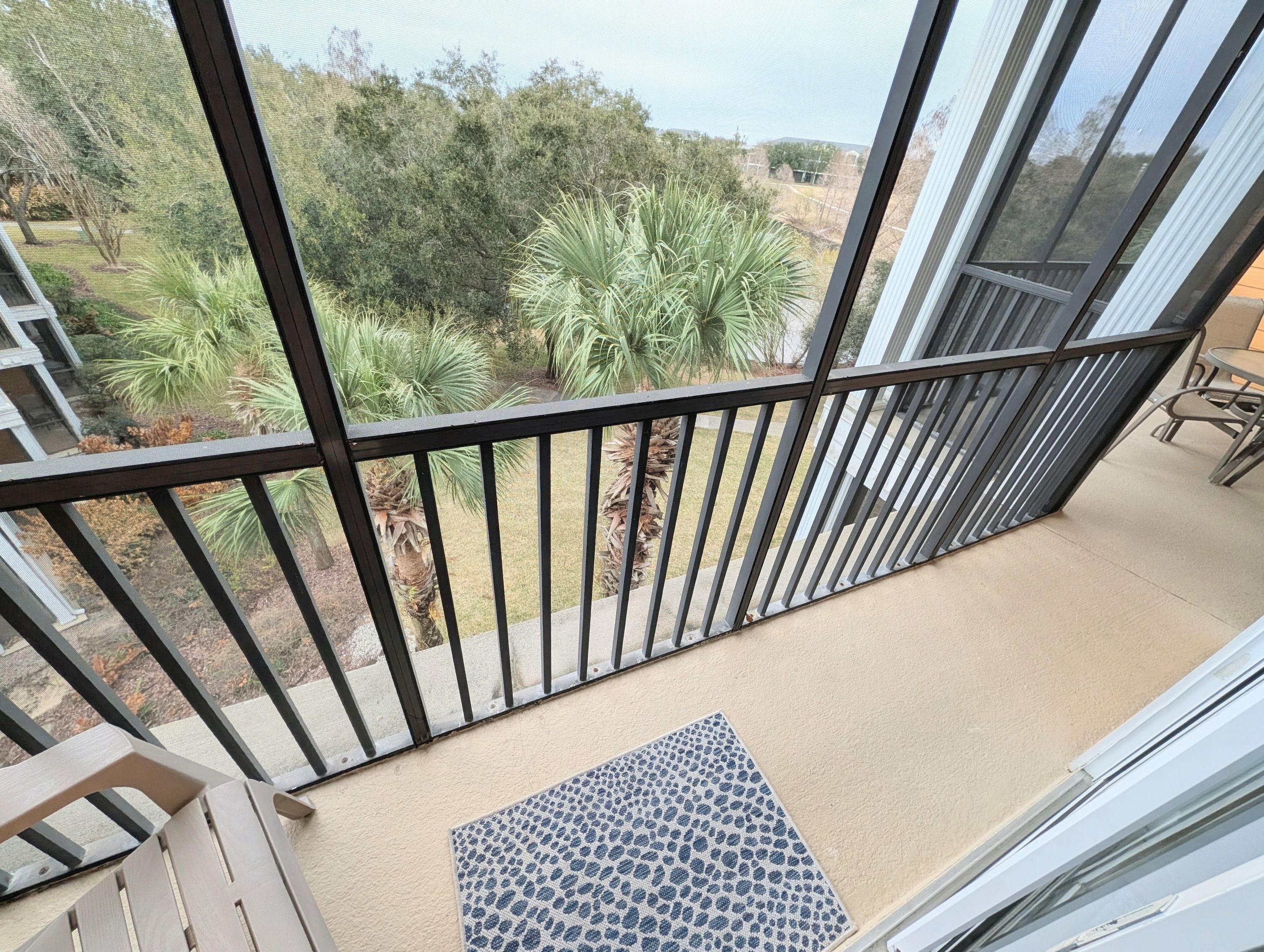 A balcony with a view of a garden, a door mat, and a table.