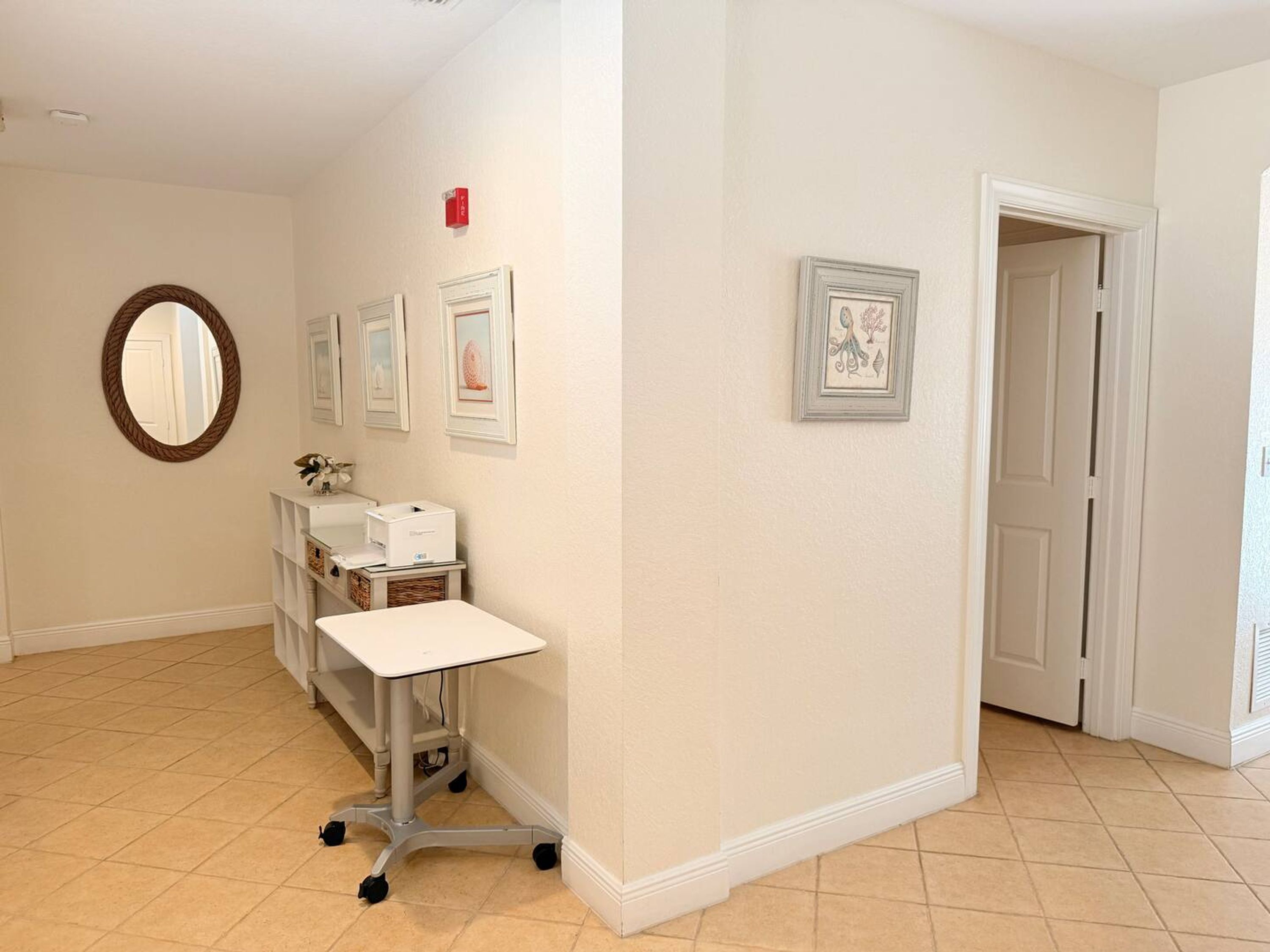 A hallway with a white tiled floor, a white desk with a computer, a mirror, and framed artwork on the walls.