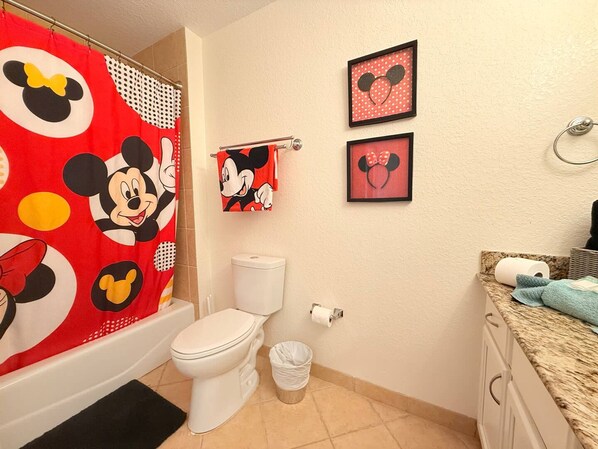 A bathroom with a Mickey Mouse shower curtain, a toilet, a trash can, and two framed Mickey Mouse head pictures on the wall.