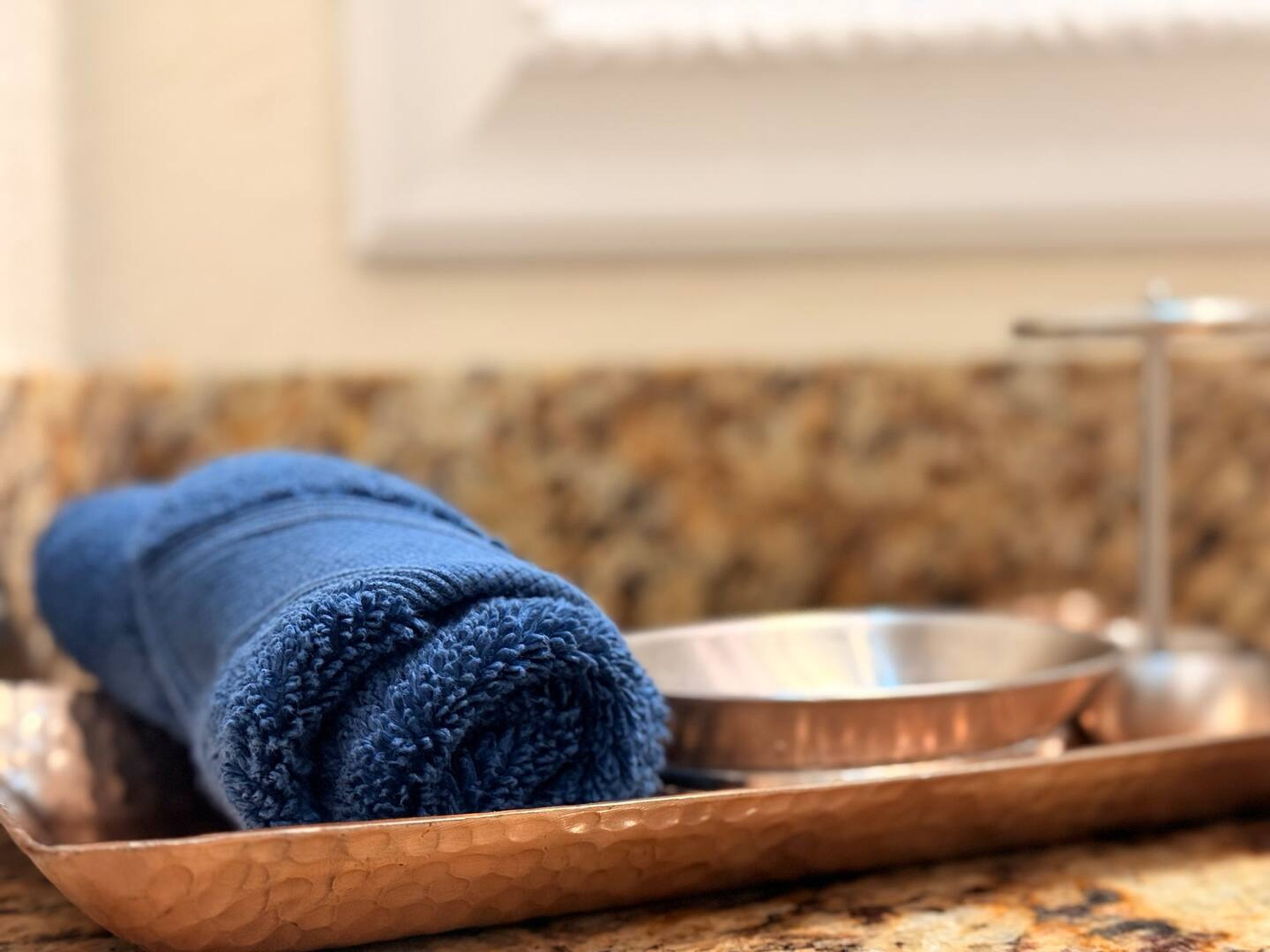 Une serviette bleue roulée sur un plateau en cuivre, un bol en métal et un robinet sur un comptoir en granit.