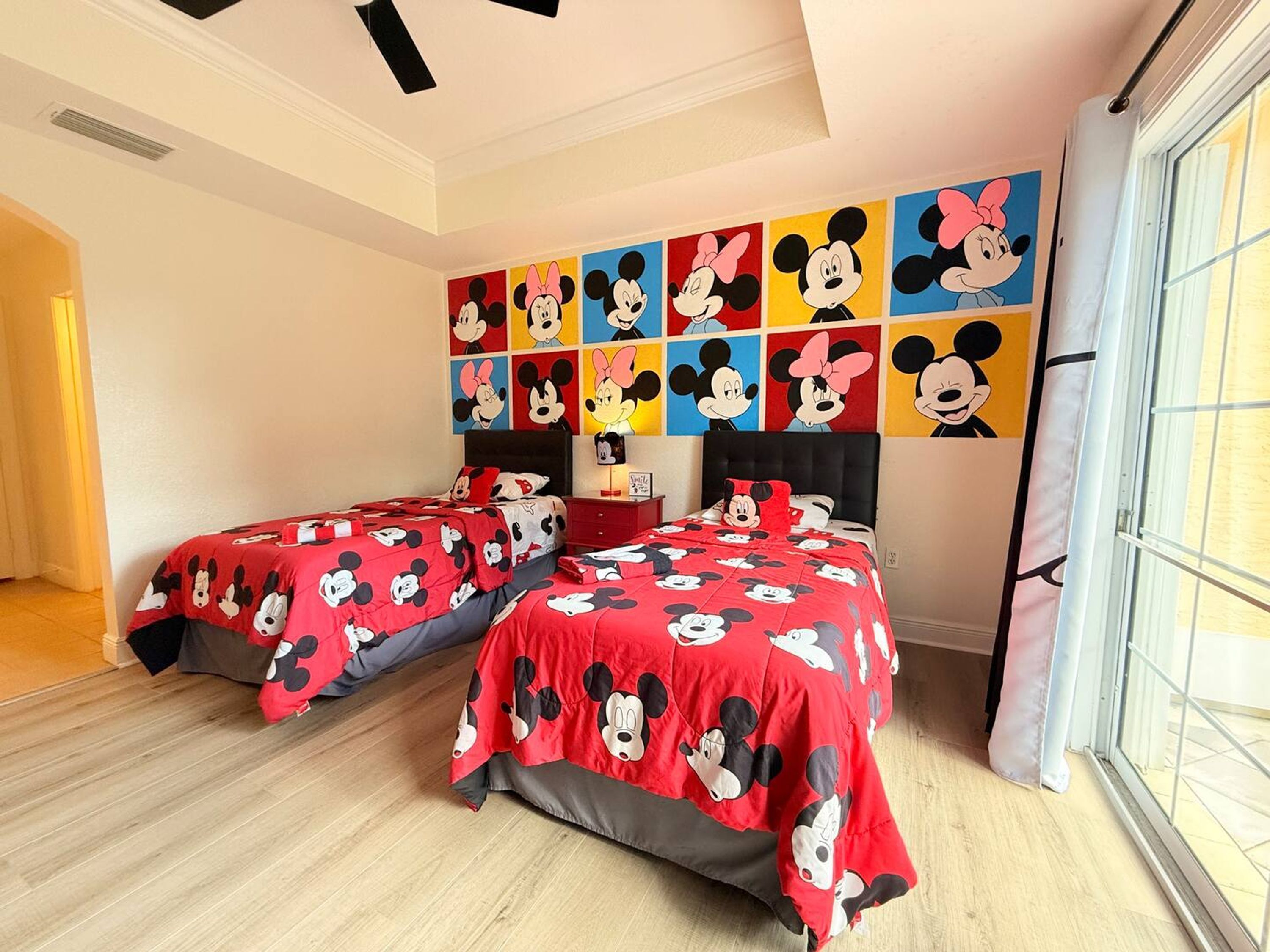 Una habitación de hotel con dos camas, cada una cubierta con ropa de cama roja con estampado de Mickey Mouse. La habitación tiene un ventanal grande en el lado derecho y un techo con vigas negras.