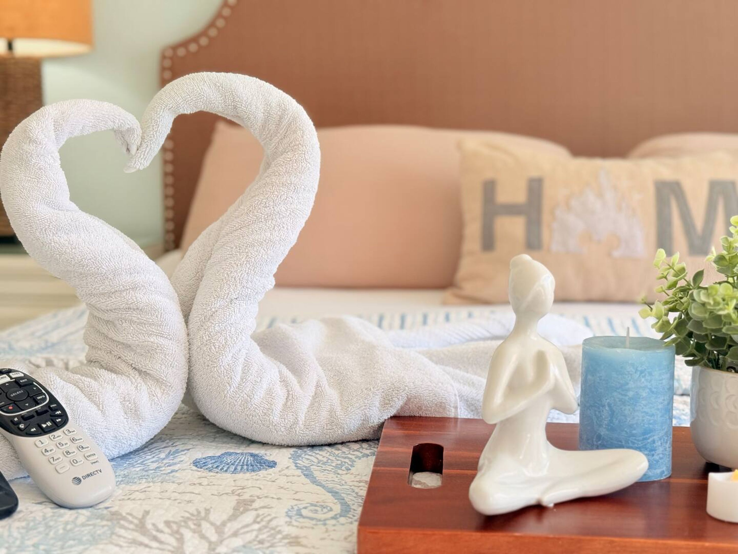 Ein Bett mit einer herzförmigen Handtuchgestaltung, einer Fernbedienung, einer kleinen Statue, einem blauen Kerzen und einem Zierkissen mit dem Buchstaben 'H'.