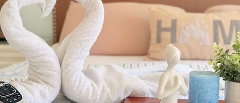 Ein Bett mit einer herzförmigen Handtuchgestaltung, einer Fernbedienung, einer kleinen Statue, einem blauen Kerzen und einem Zierkissen mit dem Buchstaben 'H'.