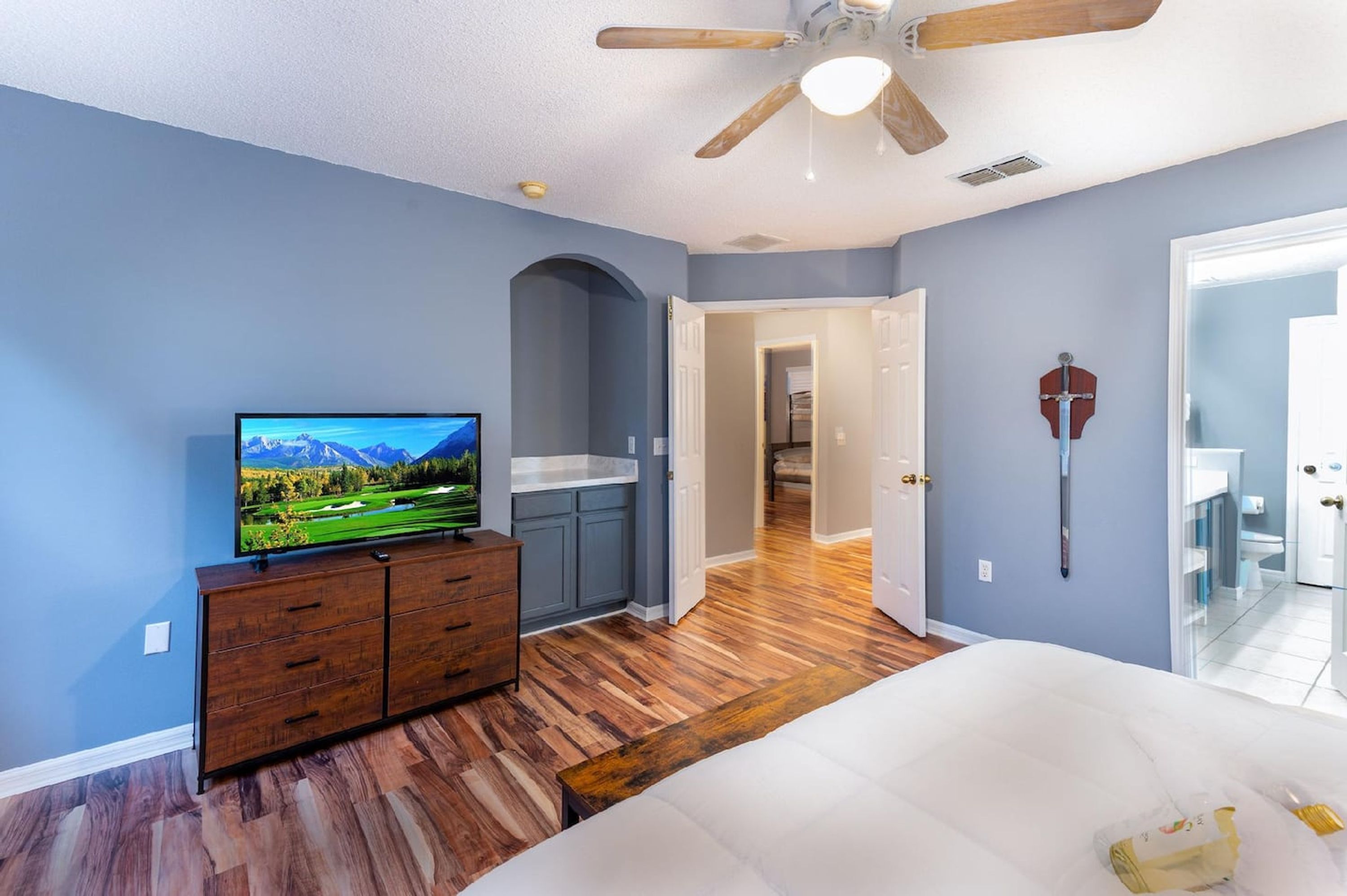 A bedroom with a bed, a TV on a wooden dresser, a ceiling fan, and a view of a bathroom.