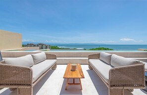 A rooftop terrace with wicker furniture, a wooden table, and a view of the ocean and distant buildings. - Rooftop Oasis in Punta Mita (Punta de Mita)