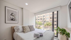 Ein modernes Schlafzimmer mit Bett, Kissen, einem gerahmten Bild und Blick auf Gebäude durch ein großes Fenster.