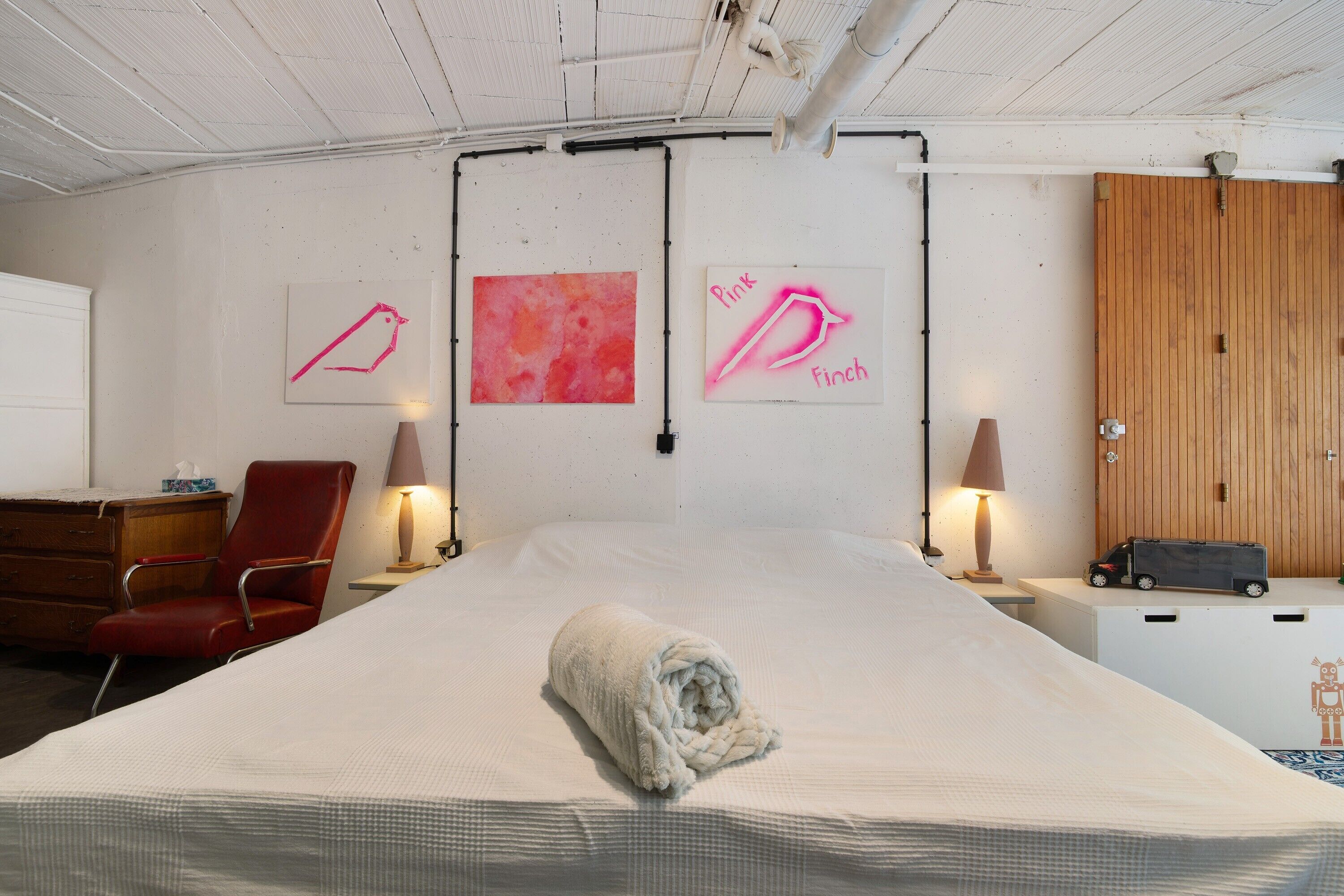 A hotel room with a large bed, a red chair, two wall-mounted lamps, and artwork depicting a pink finch.