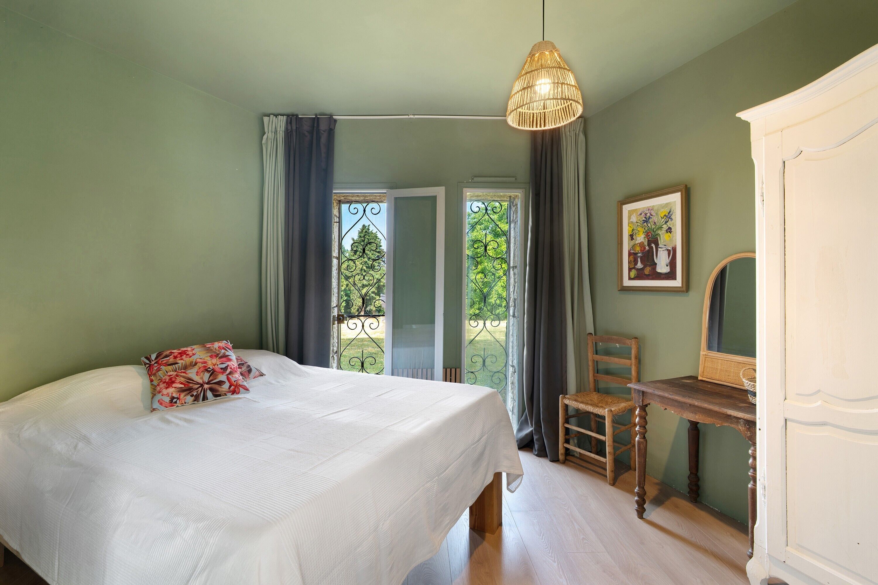 Ein Schlafzimmer mit einem Bett, einem Sessel, einer Kommode, einer Pendelleuchte und einem Fenster mit Vorhängen.