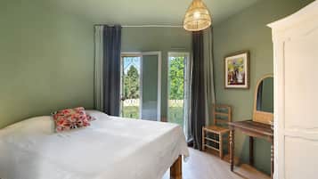 Ein Schlafzimmer mit einem Bett, einem Sessel, einer Kommode, einer Pendelleuchte und einem Fenster mit Vorhängen.