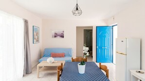 Una habitación con un sofá azul, una mesa de madera, un jarrón blanco con una planta, una puerta azul y un refrigerador blanco.
