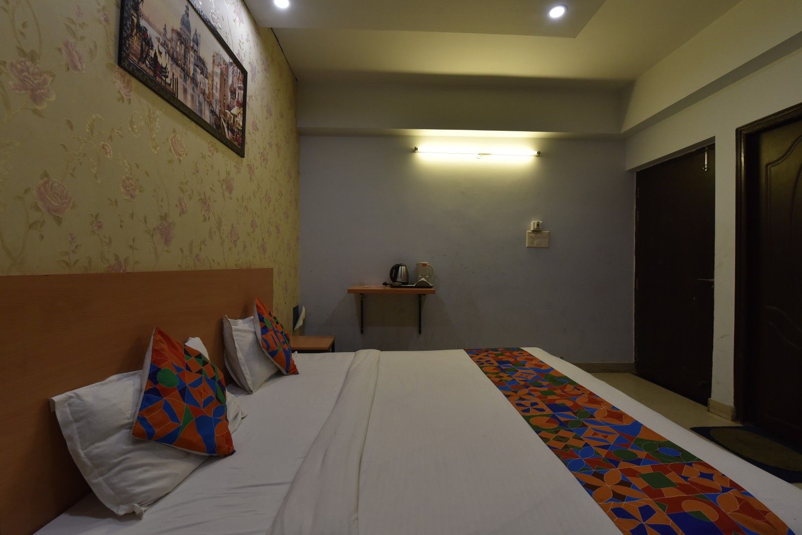 Ein Hotelzimmer mit Bett, Kissen, Nachttisch, Lampe, Wandregal, Tür und Teppich mit Muster.