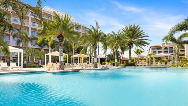 A large, clear swimming pool with lounge chairs and palm trees, surrounded by hotel buildings. - Oceanfront 2BR Condo Le Vent 309 by Bocobay (Noord)