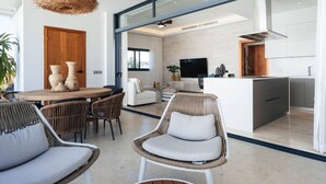 A modern interior with a dining area, wicker chairs, a kitchen with a TV, and a large window. - Penthouse 4BR w/ Pool & Sea View | Sol de Arena C7 (Las Terrenas)