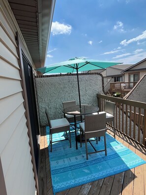 A covered outdoor patio with a table and chairs, a green umbrella, and a wooden deck. - 2BR Condo,Pools & Walk to Beach (Ocean City)