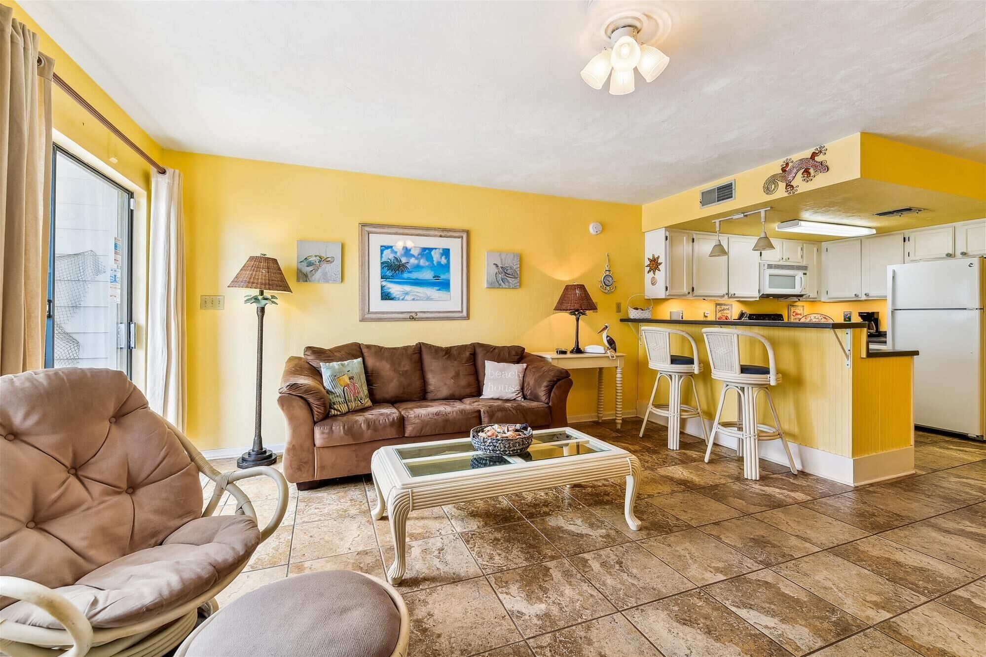 A living room with a brown leather sofa, a glass coffee table, and a wicker chair. There is a kitchen area with white cabinets and a microwave.