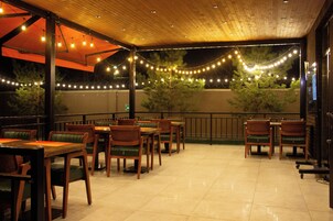 Un patio al aire libre con mesas y sillas de madera, luces colgantes y vista a árboles.