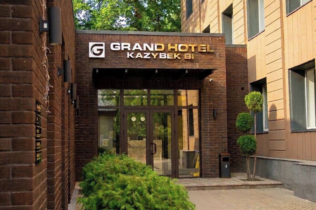 Fachada de ladrillo del Grand Hotel Kazybek B1, con puertas de entrada de vidrio y un pequeño árbol en primer plano.