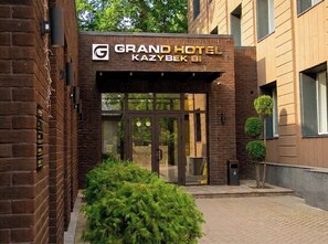 The exterior of the Grand Hotel Kazybek B1 with a brick facade, glass entrance doors, and a small tree in the foreground. - Grand Hotel Kazybek Bi (Almaty)