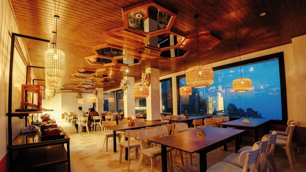 A restaurant with wooden ceiling, hexagonal light fixtures, and large windows overlooking a cityscape.
