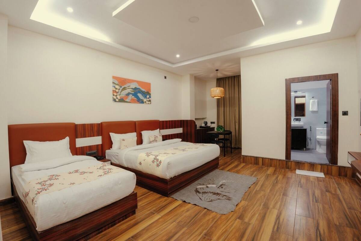 A hotel room with two beds, a desk, a chair, a small table, and a bathroom visible through an open door.
