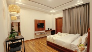 A hotel room with a wooden floor, a bed with white bedding, a TV mounted on the wall, a desk with a chair, and a large window with curtains.