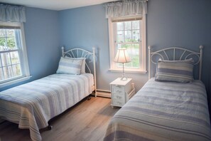 A bedroom with two single beds, a nightstand, a lamp, and a window with a view of greenery. - Charming 3 bedroom home, .5 miles to Thatcher Beach. (South Yarmouth)