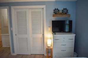 A room with a closed white door, a television on a dresser, a small table with a lamp, and a wall-mounted shelf with nautical-themed decor. - Charming 3 bedroom home, .5 miles to Thatcher Beach. (South Yarmouth)