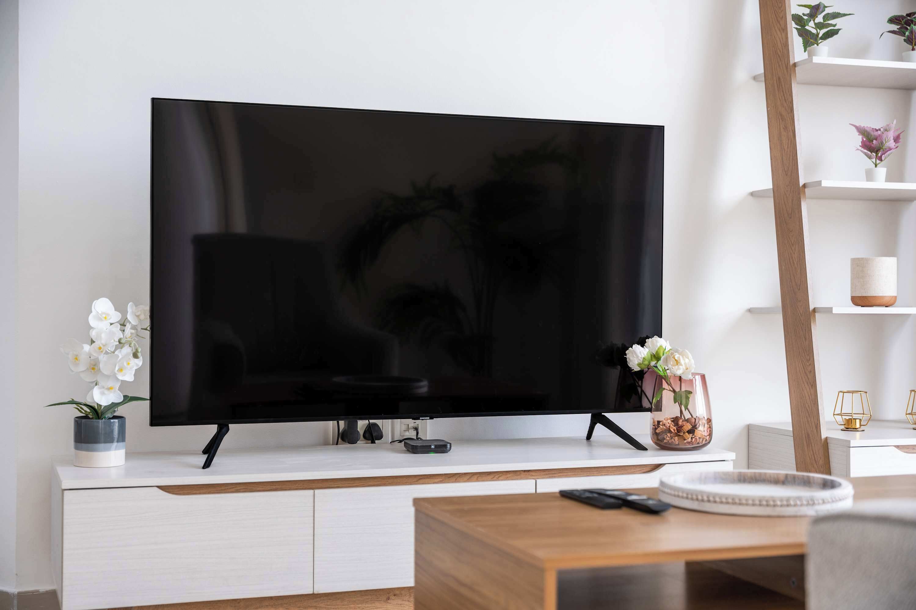 Una TV a schermo piatto montata su un mobile bianco, un tavolino da caffè in legno in primo piano e una mensola con oggetti decorativi.