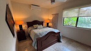 Quarto com cama, duas mesas de cabeceira com luminárias, ventilador de teto, ar condicionado, janela com vista para vegetação e uma obra de arte na parede.