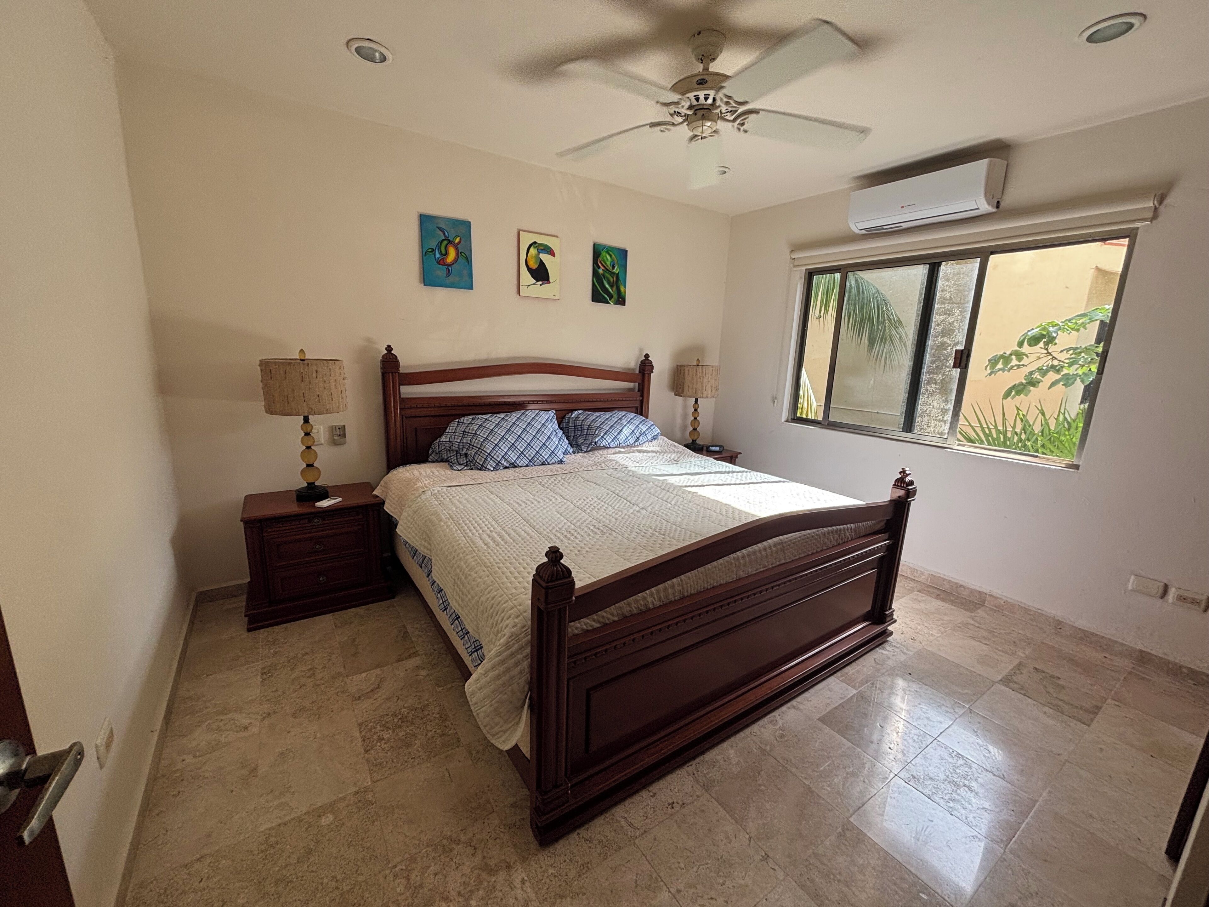 Un dormitorio con una cama de madera, dos mesitas de noche con lámparas, un ventilador de techo y una ventana con vistas a la vegetación.