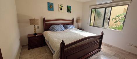Un dormitorio con una cama de madera, dos mesitas de noche con lámparas, un ventilador de techo y una ventana con vistas a la vegetación.