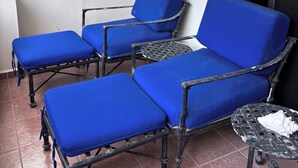 Uma área de pátio com cadeiras azuis acolchoadas e uma mesa pequena, uma luminária na parede e uma janela com vista para o verde.