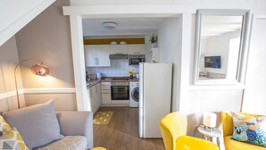 A compact living space with a kitchenette, a sofa, a yellow armchair, and a small table with a lamp. - Poachers Cottage - 3 Bedroom Cottage - Ivy Towers (Tenby)