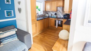 A compact kitchen with wooden cabinets, a black countertop, and a window with a view of greenery. - Old Tom Cottage - 3 Bedroom House - St Florence (Tenby)