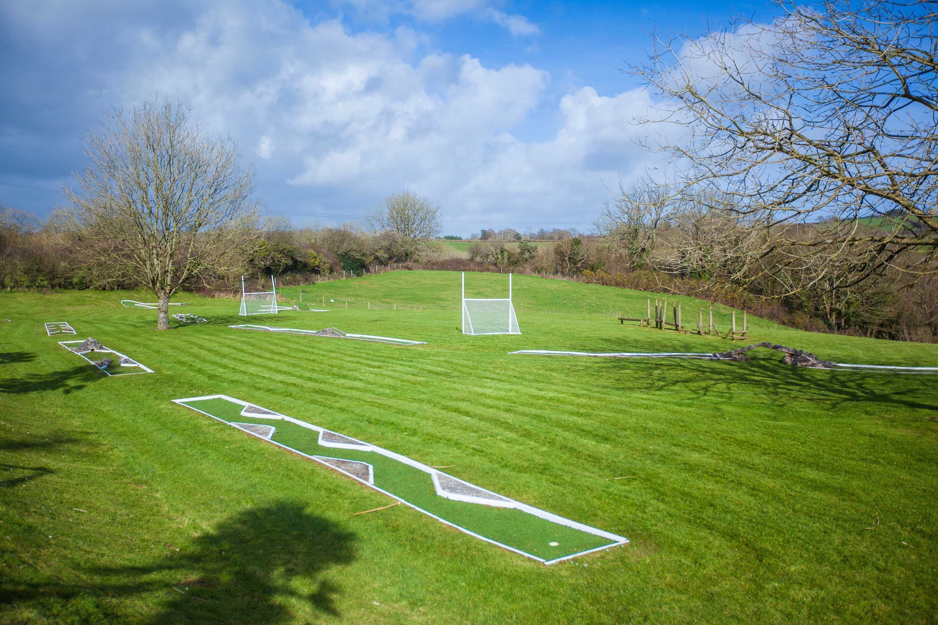 A well-maintained grassy field with goalposts and a soccer goal, surrounded by trees and open sky.