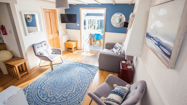 A living room with a blue patterned rug, a grey sofa, a wooden side table, a white armchair, and a television mounted on the wall. - Old Tom Cottage - 3 Bedroom House - St Florence (Tenby)