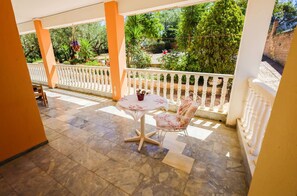 A covered outdoor area with a table and chair, a white railing, and a view of greenery.