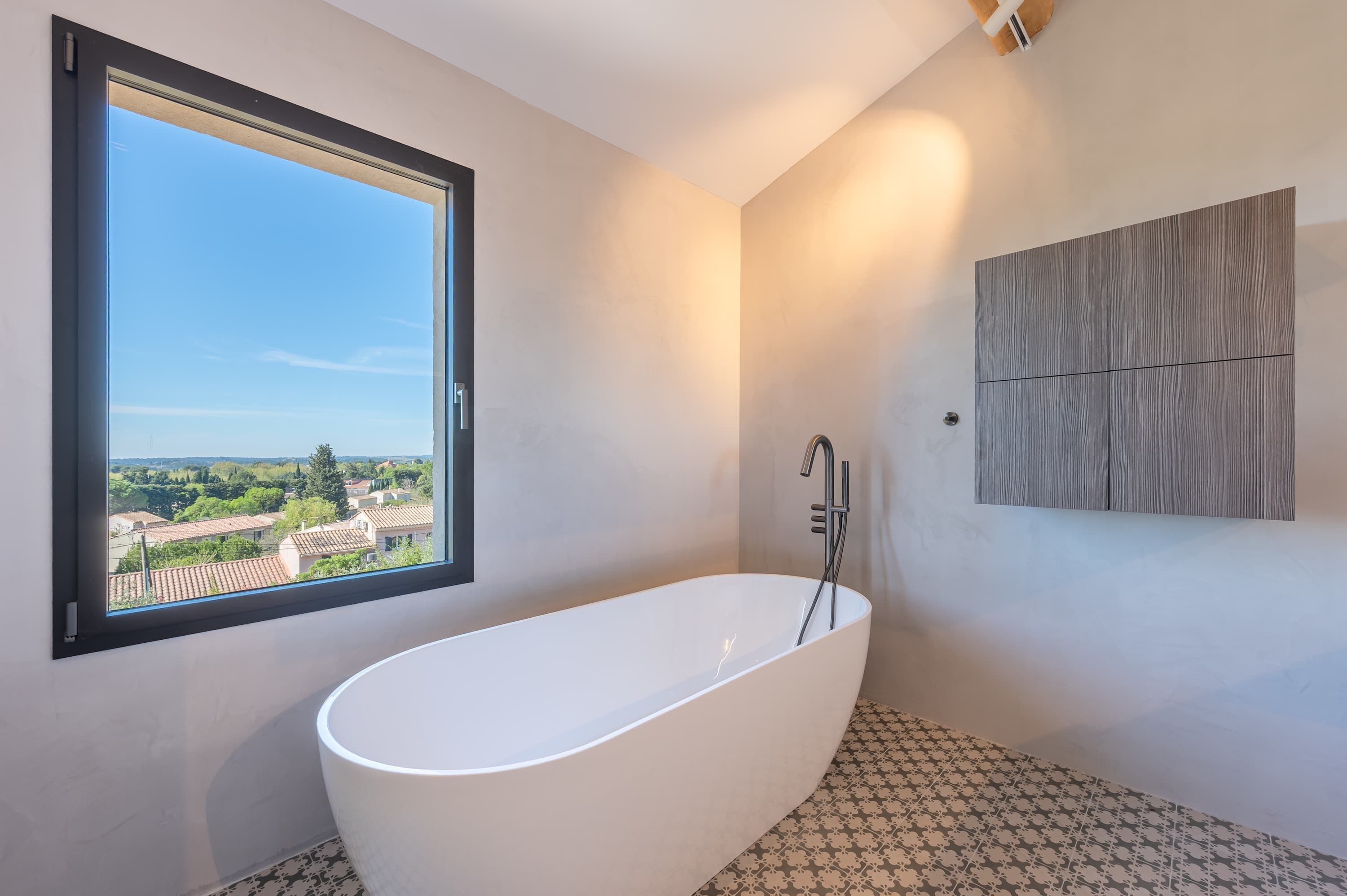 Une salle de bain moderne avec une baignoire sur pieds, une grande fenêtre offrant une vue sur les bâtiments et la verdure, et un panneau en bois fixé au mur.