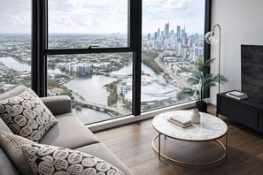 A modern living room with a large window, a sofa, a round coffee table, a potted plant, and a TV stand. - 2BR Resort Style Apartment with Panoramic Views (Broadbeach)