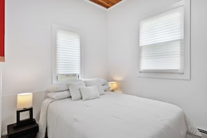 Une chambre à coucher avec un lit, deux tables de chevet et deux fenêtres équipées de stores.