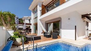 A modern poolside area with a clear view of the pool, outdoor furniture, and a building with large windows. - La Manga Club Atalayas II Villa 616 (Cartagena)