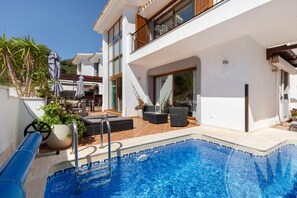 Un área moderna junto a la piscina con vista despejada, mobiliario exterior y un edificio con grandes ventanales.