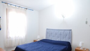 A bedroom with a bed, a nightstand with a lamp, a wooden table, and a window with curtains. - Apartment Faro in Budoni (Budoni)
