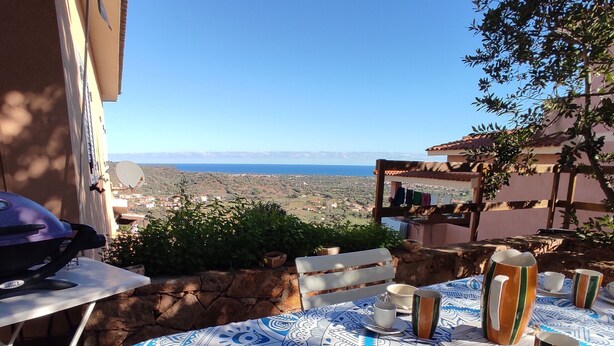 An outdoor dining area with a view of the sea, a table set with plates, cups, and a pitcher, and a grill on the left. - Apartment Faro in Budoni (Budoni)