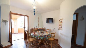 A dining area with a round table, chairs, a chandelier, a TV mounted on the wall, and a brick fireplace. - Apartment Faro in Budoni (Budoni)