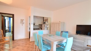 A dining area with a table and chairs, a kitchen area with cabinets and appliances, and a television on a stand. - House Gavina 2 Seaside Alghero (Fertilia)