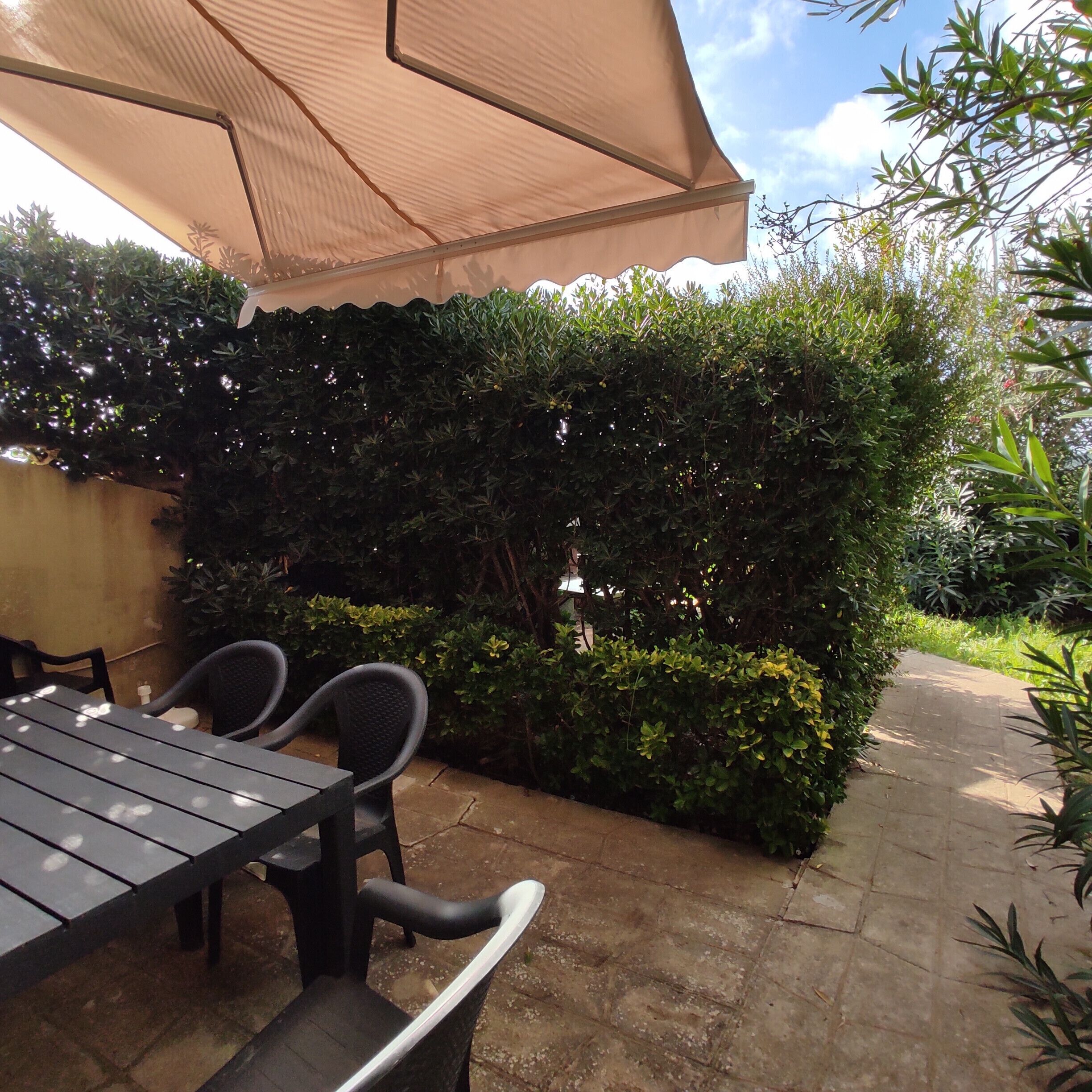 An outdoor patio with a table and chairs, a large umbrella, and a hedge.