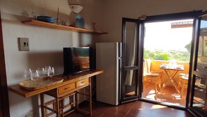 Ein kompakter Wohnbereich mit Schreibtisch aus Holz, einem Fernseher und einem Kühlschrank. Eine Glastür öffnet sich zu einer Außenveranda mit Tisch und Stühlen.